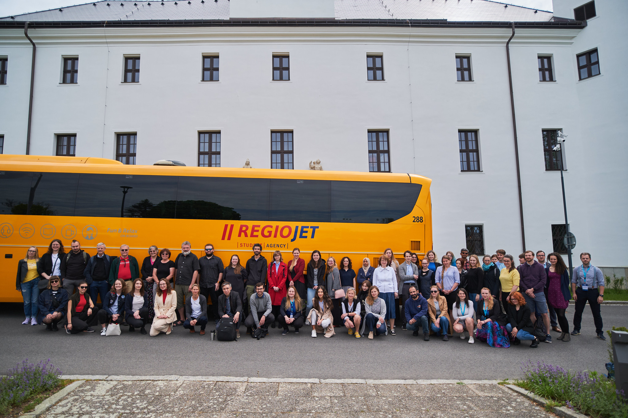 Skupinová fotografie účastníků konference před žlutým autobusem společnosti RegioJet. V pozadí budova Zámku Třešť.