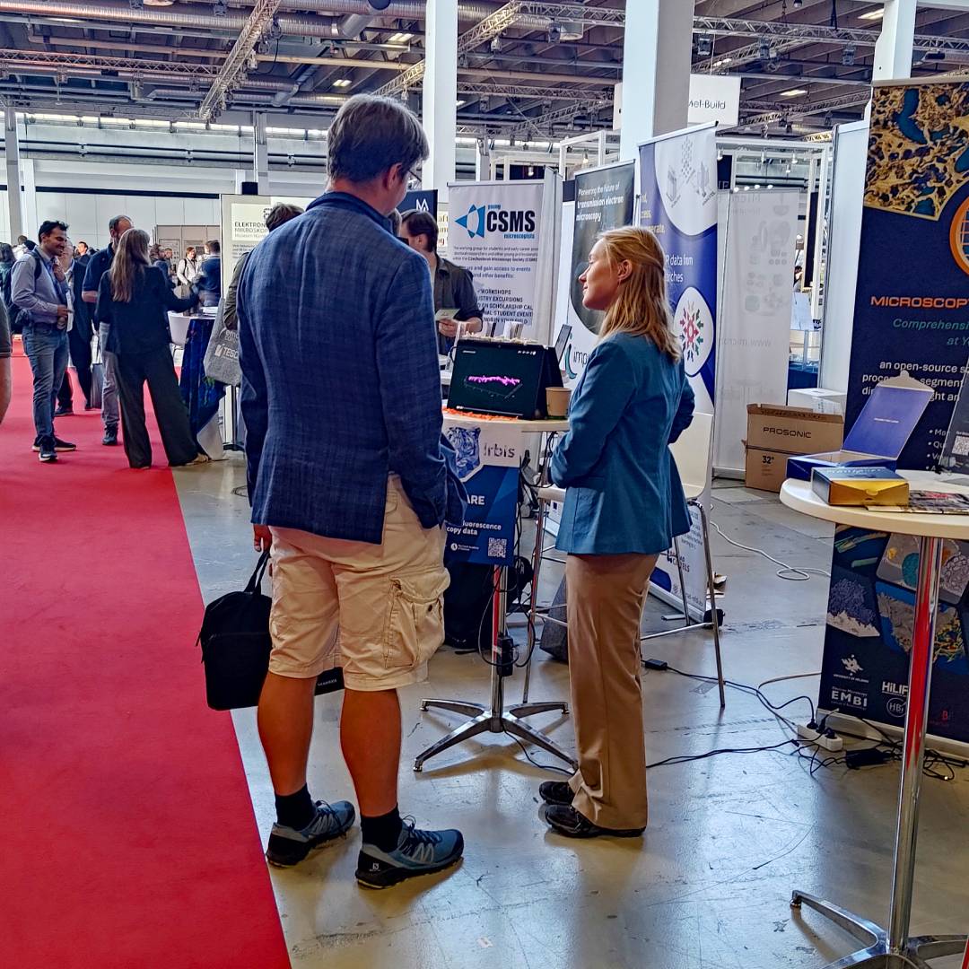 View of the IEM CAS exhibition stand with a height advertising banner and visitors discussing with scientist Iva Švecová.